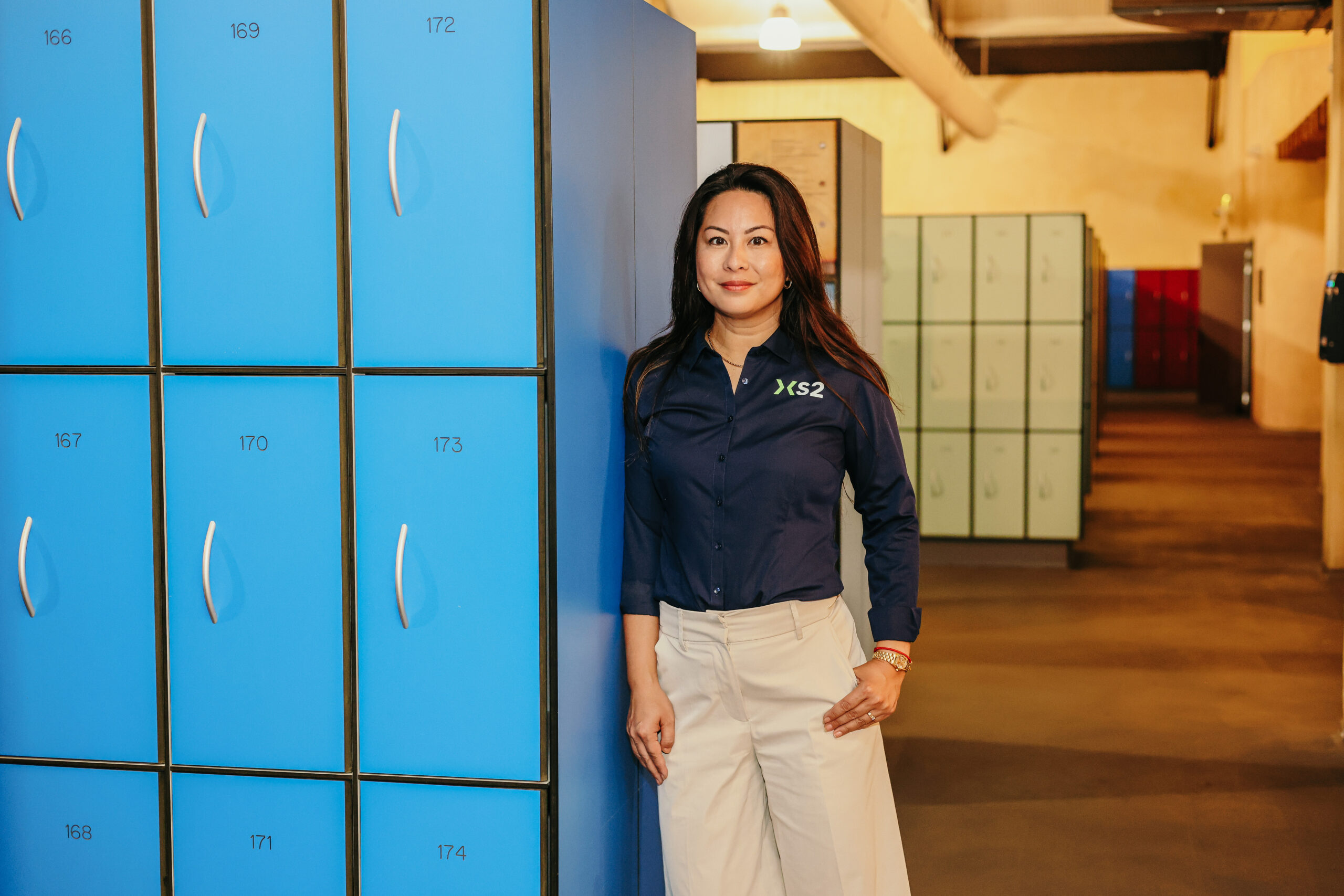 Vrouw in XS2-bedrijfskleding poseert voor blauwe lockers.