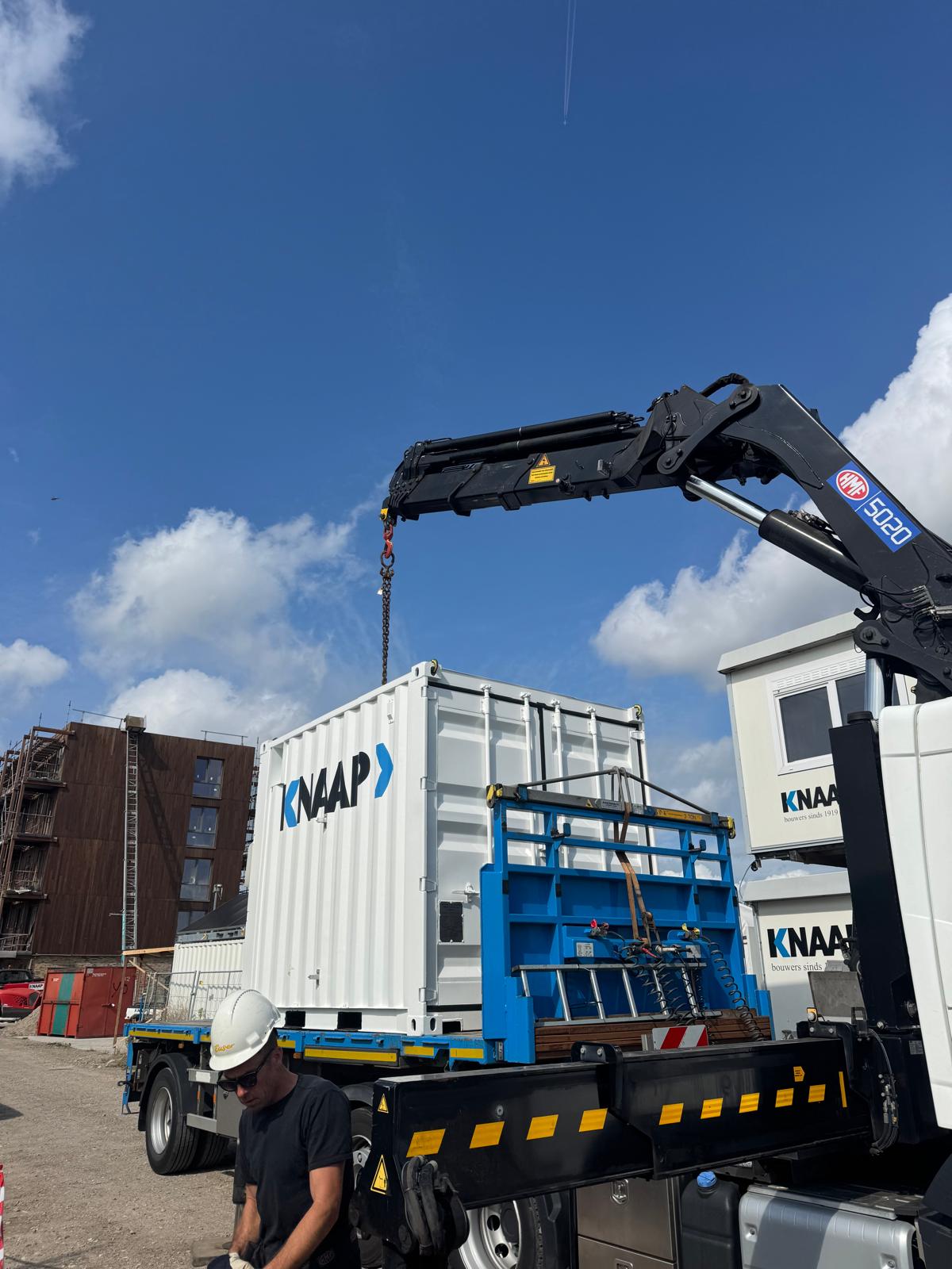 Hijskraan plaatst witte Knaap-container op een vrachtwagen op een bouwterrein.