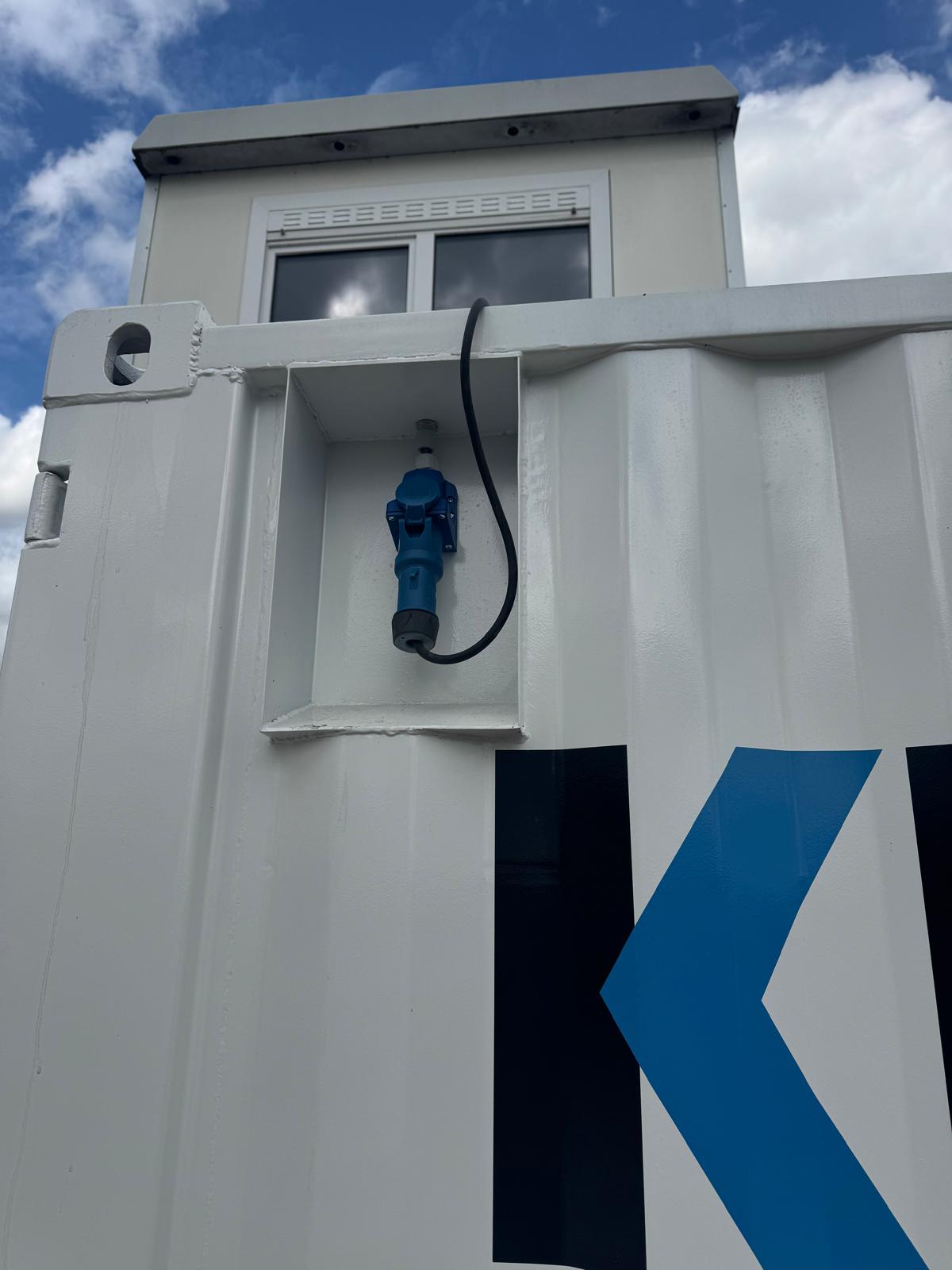 Stroomaansluiting met blauwe stekker op witte containerwand onder een raam.