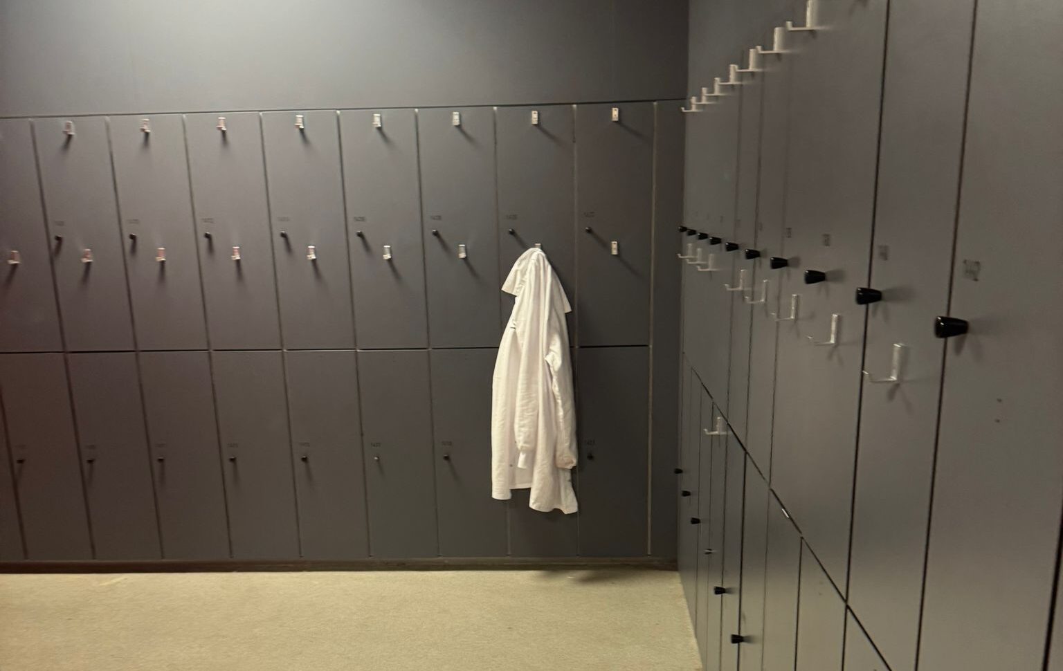 Grijze kleedruimte met meerdere rijen kluisjes en haken, waaraan een witte jas hangt.