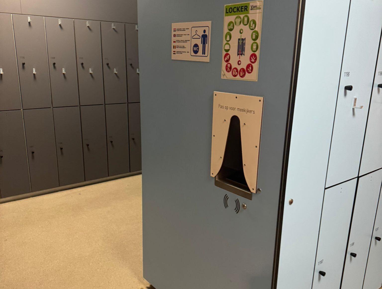 Kleedruimte met grijze en lichtblauwe kluisjes; op de voorgrond een bedieningspaneel met afscherming en de tekst ‘Pas op voor meekijkers’, naast instructieborden voor gebruik van de lockers.