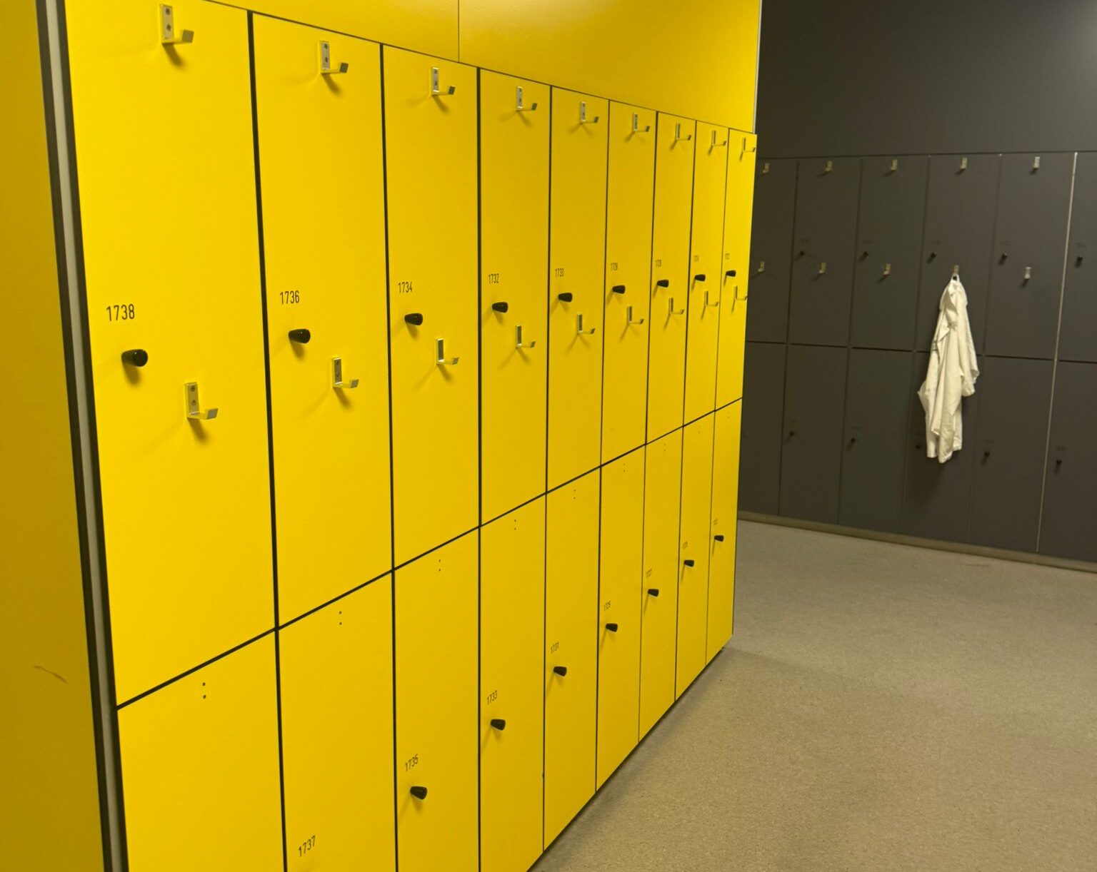 Rij felgele kluisjes met nummers in een kleedruimte, met aan de achterkant een muur met grijze kluisjes waar een witte jas aan een haak hangt.