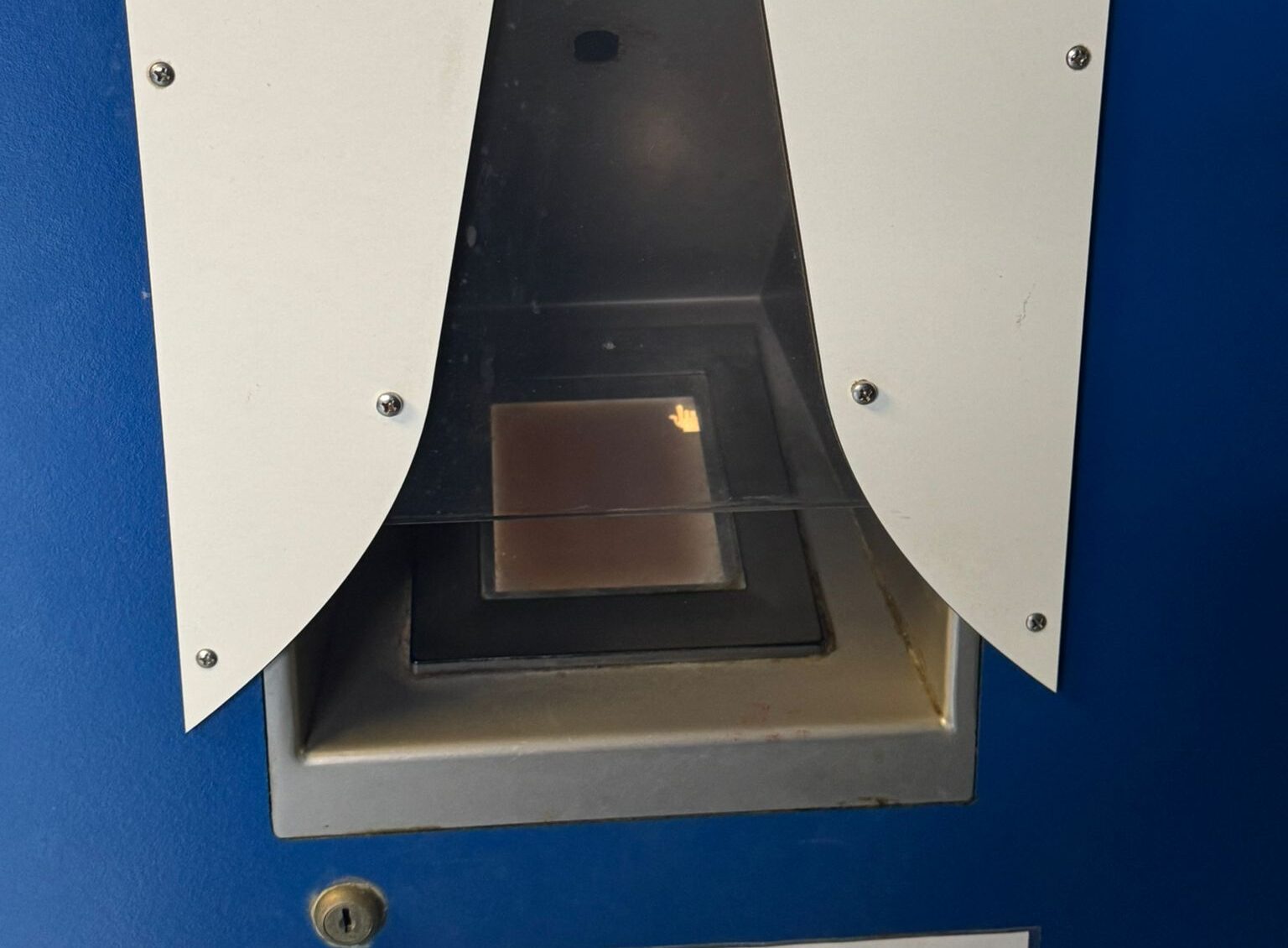 Close-up van een apparaat met een glazen plaat en een klein schermpje met een handicoon, waarschijnlijk een scanner of identificatiesysteem.