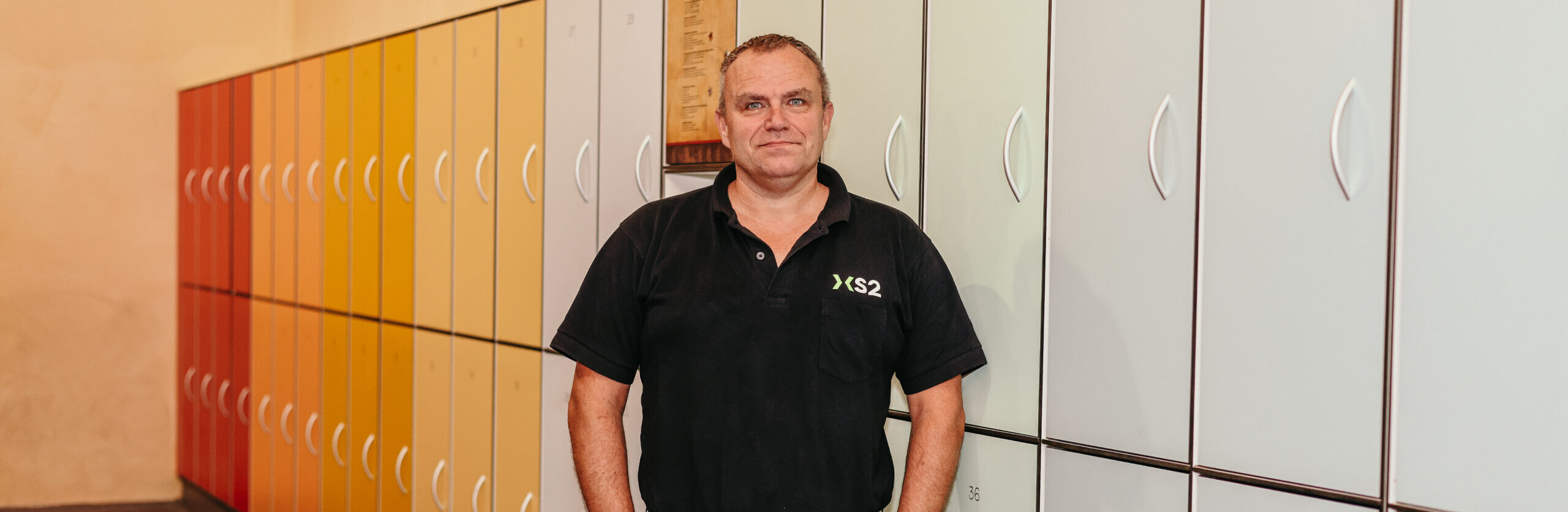 Man in XS2-shirt poseert voor een rij gekleurde lockers. Man in XS2-shirt poseert voor een rij gekleurde lockers.