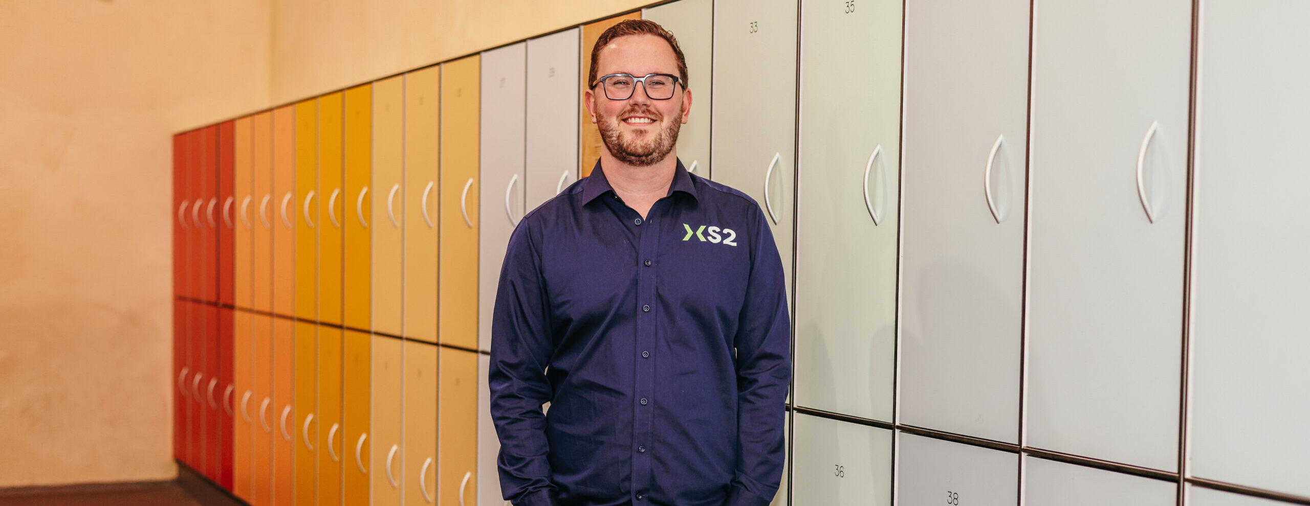 Man in XS2-bedrijfskleding staat lachend voor gekleurde lockers. Man in XS2-bedrijfskleding staat lachend voor gekleurde lockers.