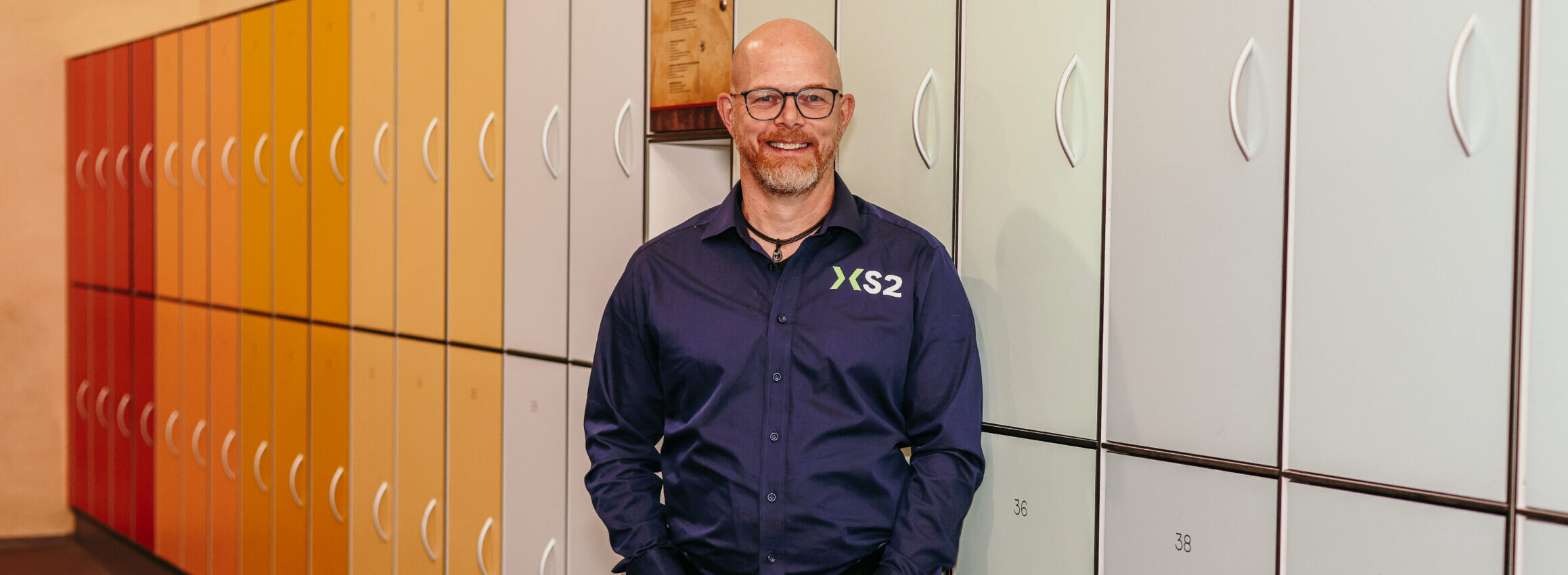 Man in XS2-bedrijfskleding staat lachend voor gekleurde lockers. Man in XS2-bedrijfskleding staat lachend voor gekleurde lockers.