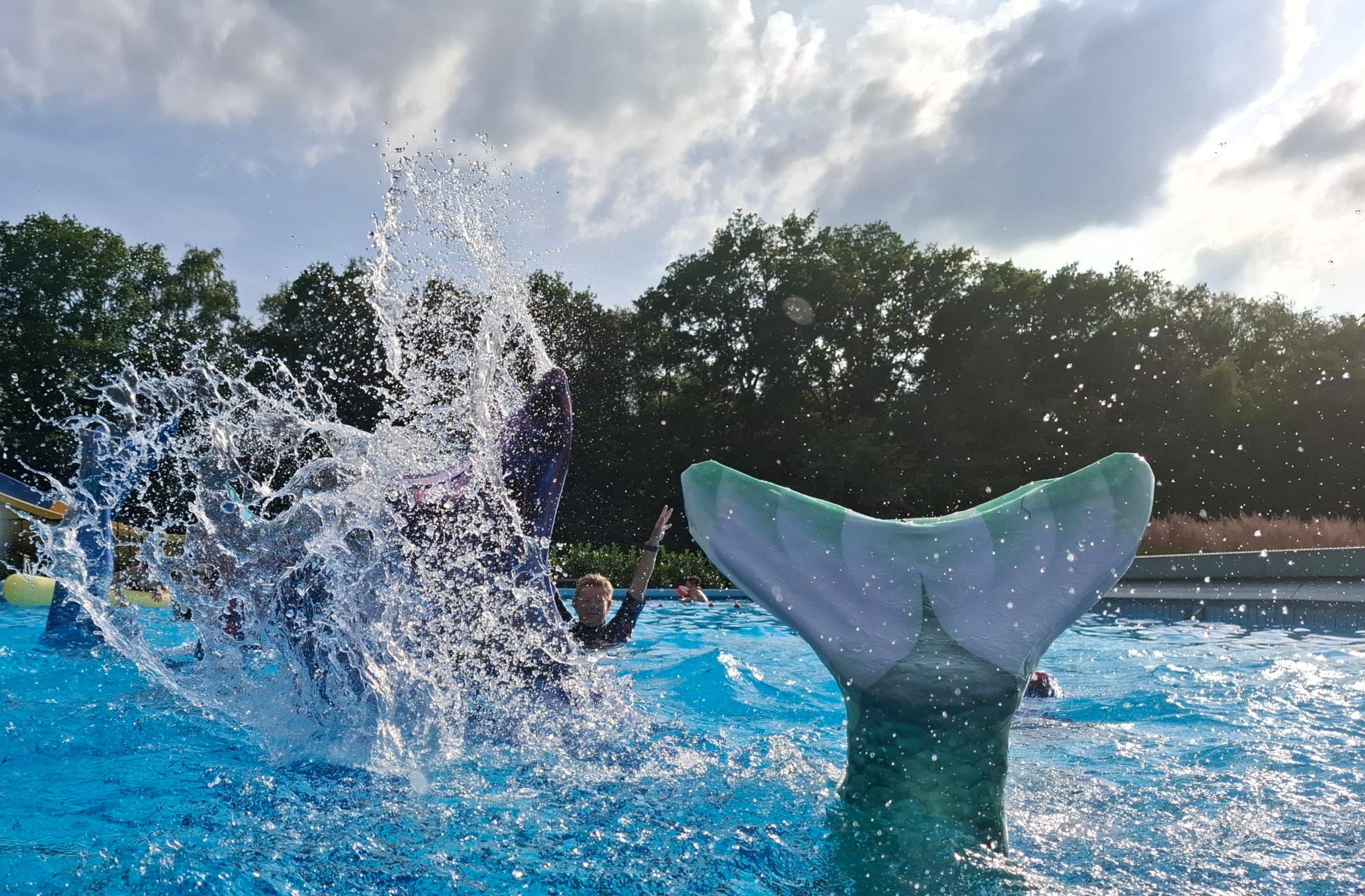 Kinderen spelen in zwembad met zeemeerminstaarten en opspattend water.
