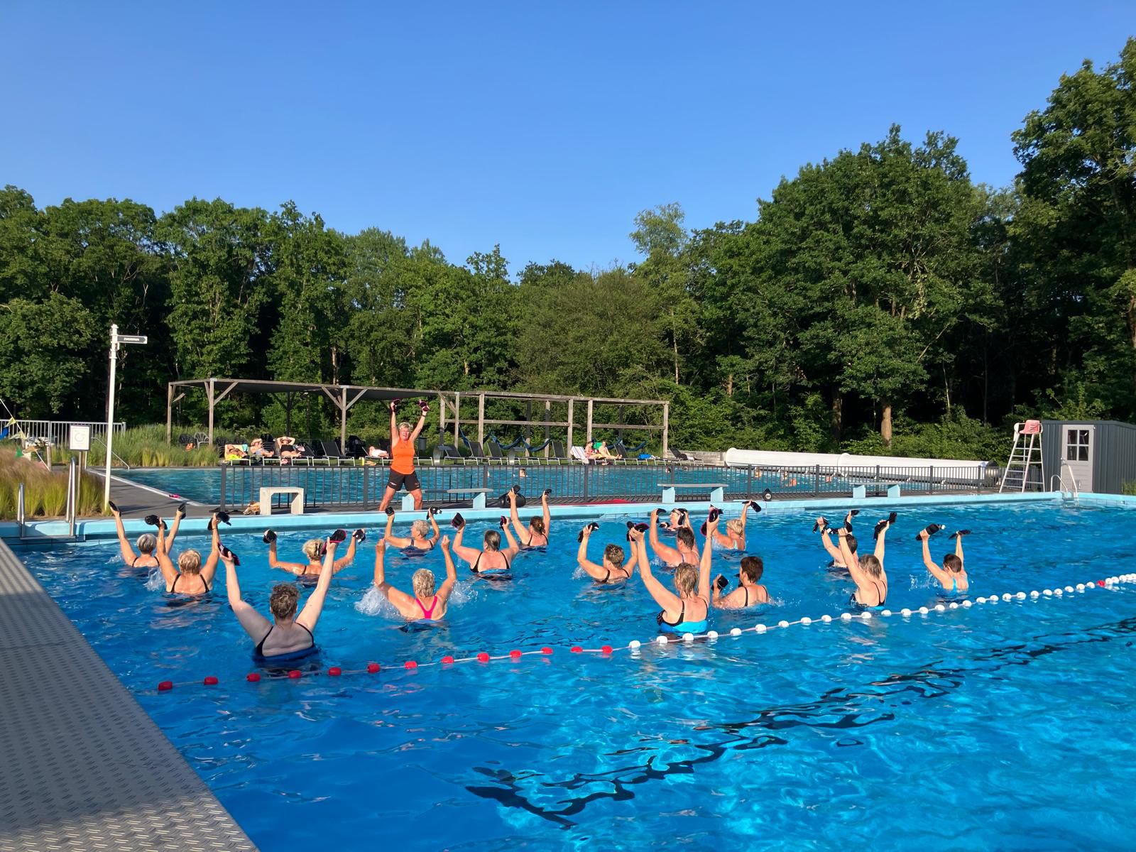 Groep mensen doet aquafitness in een buitenzwembad onder begeleiding van een instructeur.