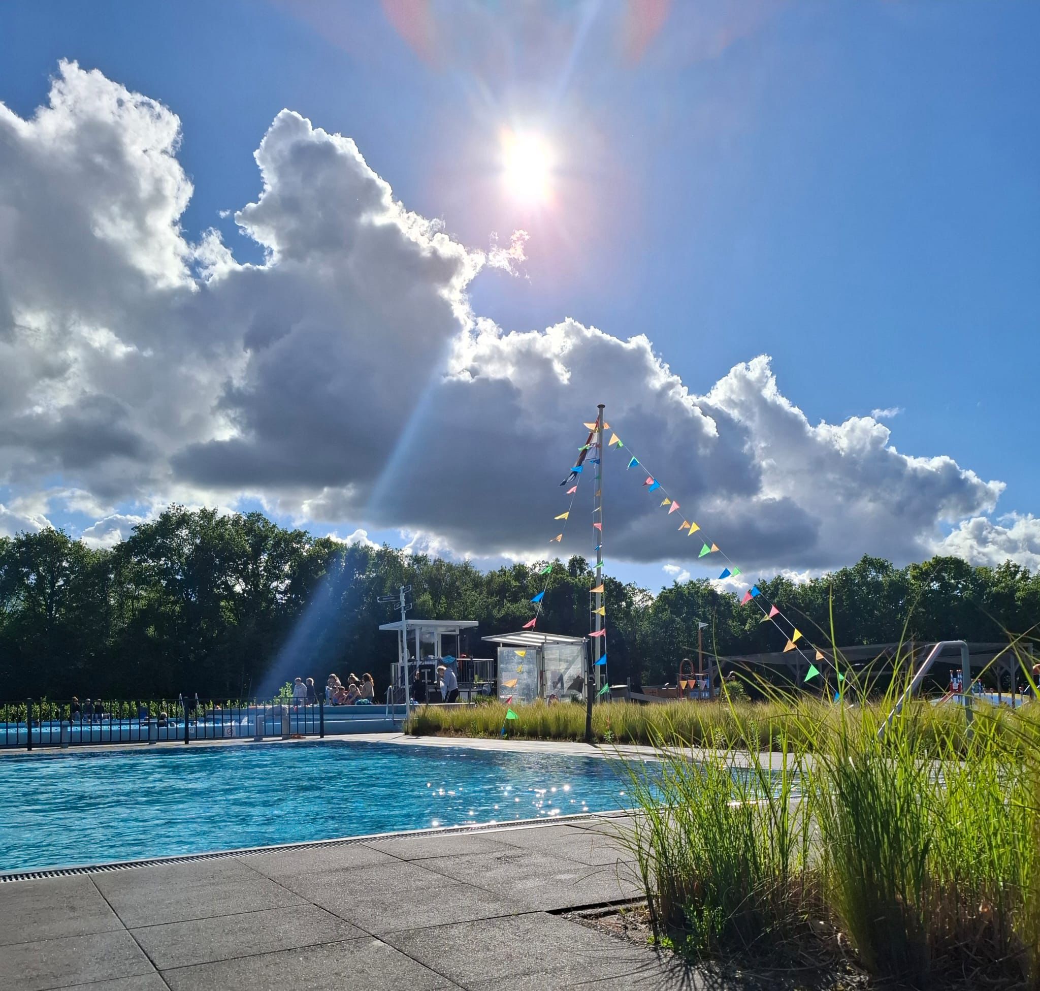 Buitenzwembad met helderblauw water, feestelijke vlaggetjes aan een paal en de zon die door de wolken straalt.