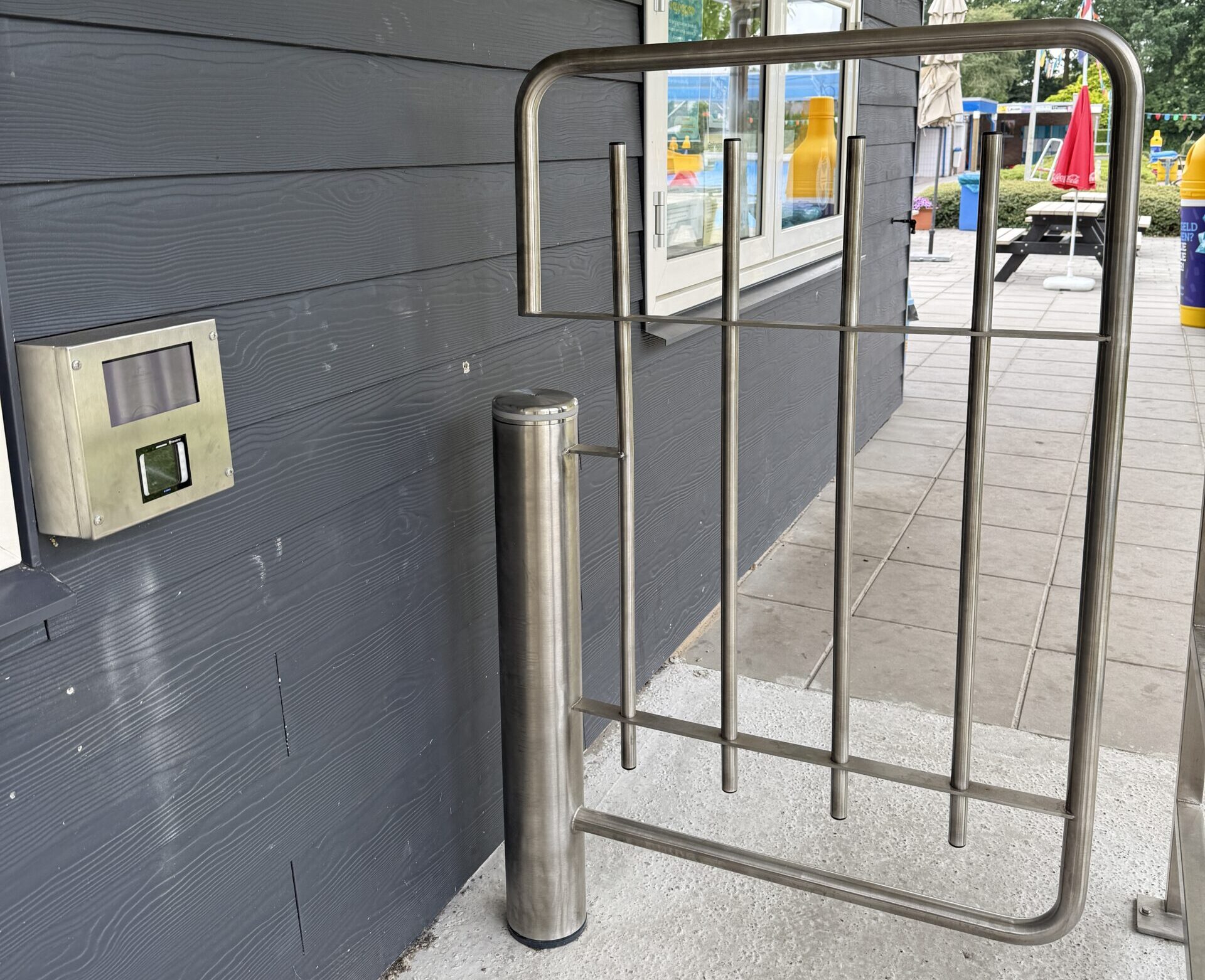 Metalen toegangshek naast een gebouw met een toegangspaneel aan de muur, op de achtergrond picknicktafels en een buitenterrein met zwembad.