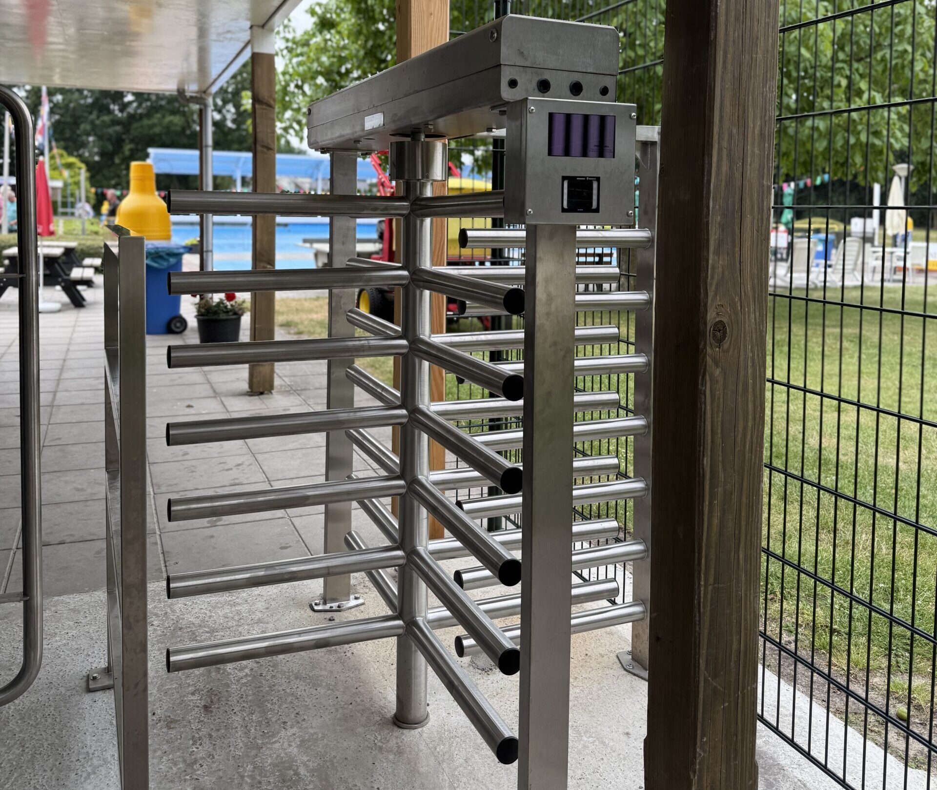 Close-up van een metalen draaipoort met toegangssysteem bij een ingang, met op de achtergrond een zwembad en buitenterrein.