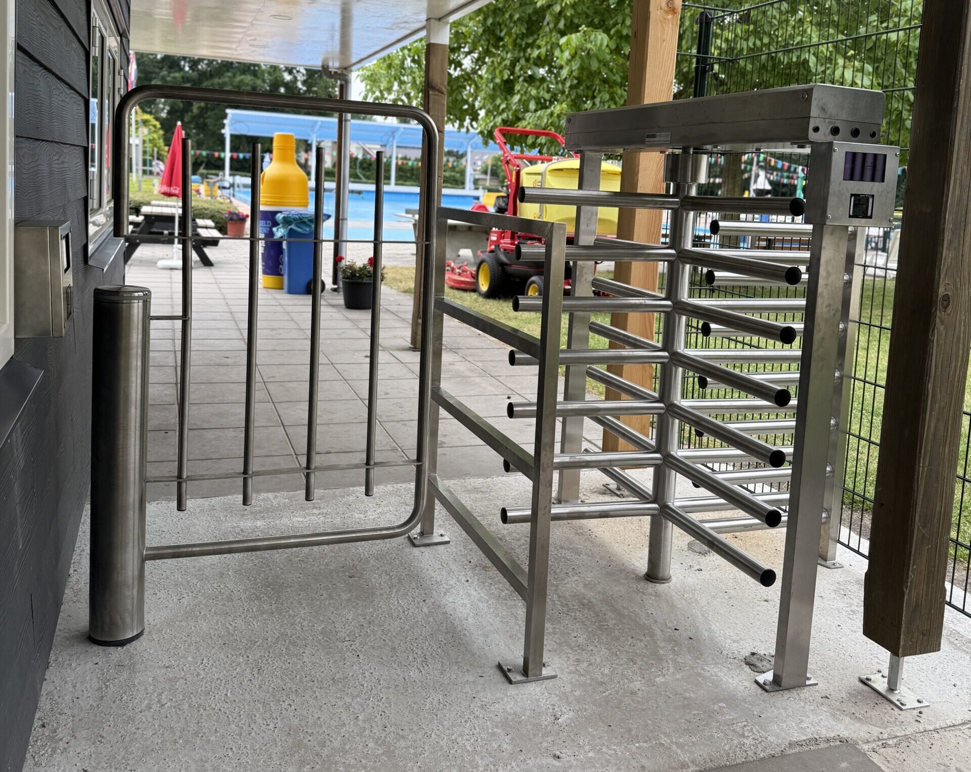 Ingang met een roestvrijstalen draaipoort en hekwerk, met op de achtergrond een zwembad, picknicktafels en kleurrijke attributen.