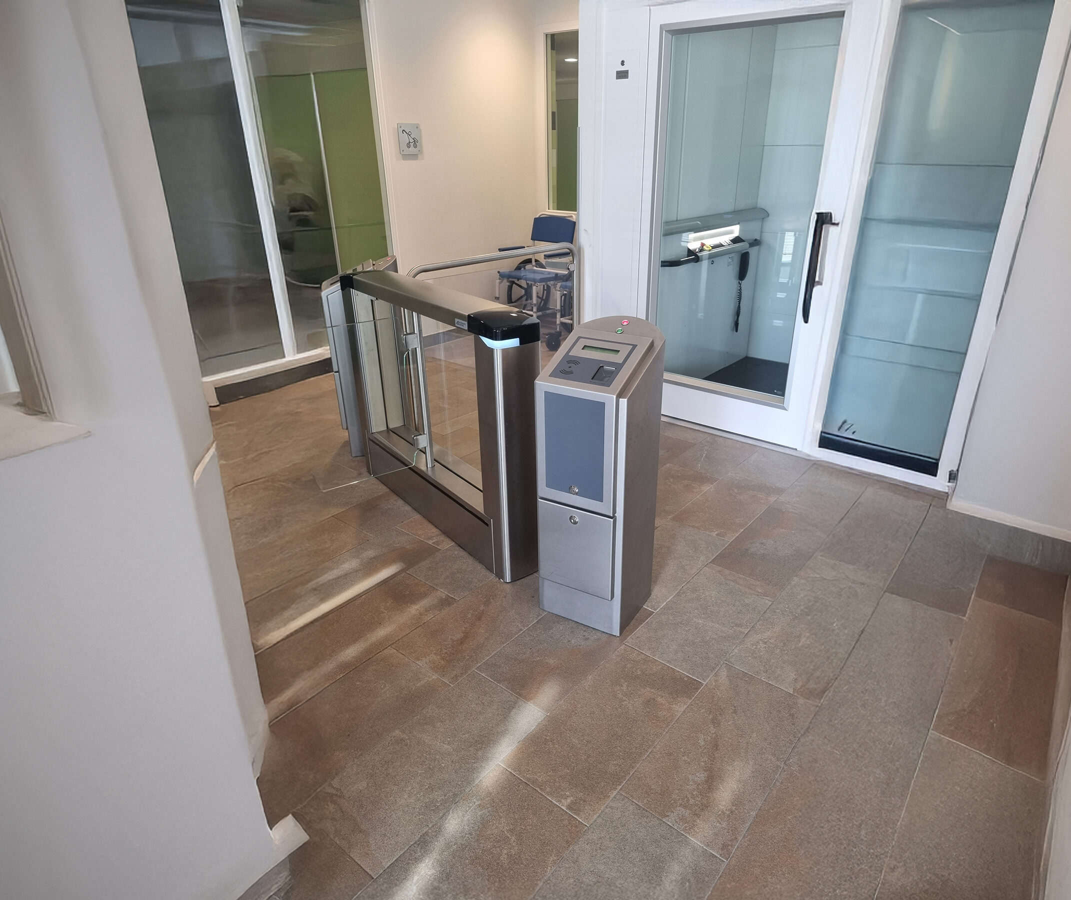 Binnenruimte met een moderne toegangspoort met glazen vleugels en een bedieningspaneel, naast een glazen liftdeur en een rolstoel.