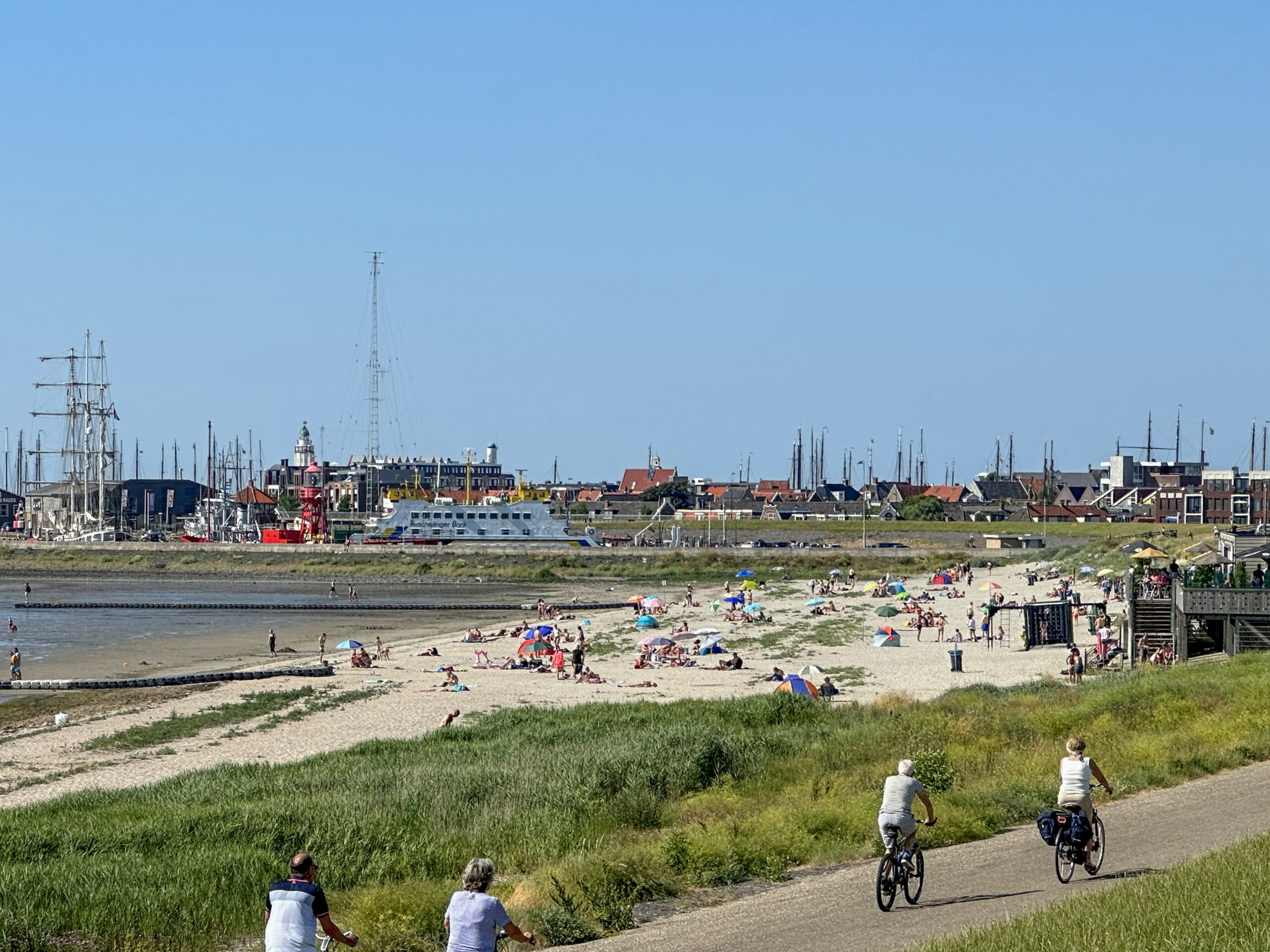 Zonnig strand met badgasten en parasols, met op de achtergrond een haven vol zeilschepen en vissersboten en op de voorgrond drie mensen die langs de kust fietsen.