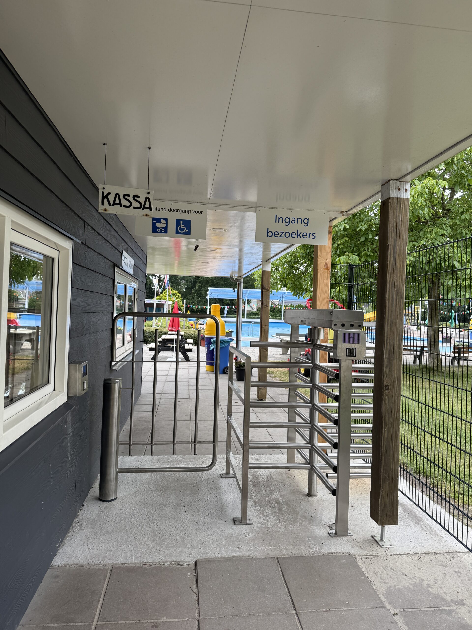 Ingang van een zwembad met draaipoort en hek, naast een kassaraam met borden ‘Kassa’ en ‘Ingang bezoekers’. Ingang van een zwembad met draaipoort en hek, naast een kassaraam met borden ‘Kassa’ en ‘Ingang bezoekers’.