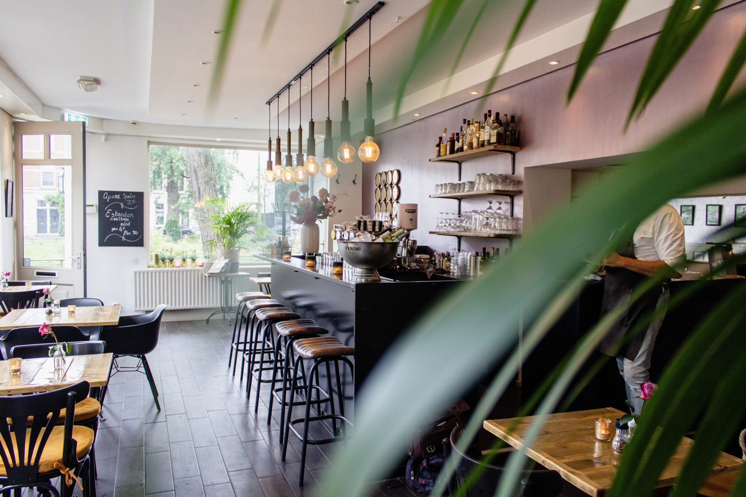 Gezellig café-interieur met houten tafels, zwarte stoelen, een bar met barkrukken en hangende lampen, gezien door bladeren van een plant op de voorgrond.